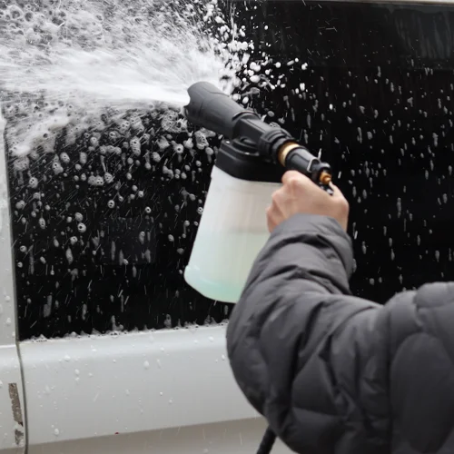 A foam gun attached to a green garden hose, spraying thick foam onto a car